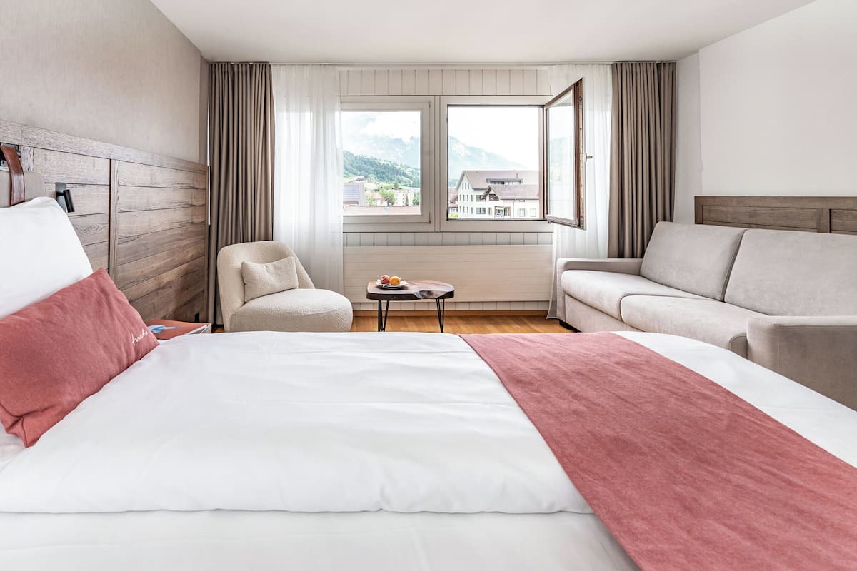 Une chambre d’hôtel avec un grand lit, deux fauteuils, une petite table avec des fruits, et une vue sur les bâtiments et les montagnes par la fenêtre.
