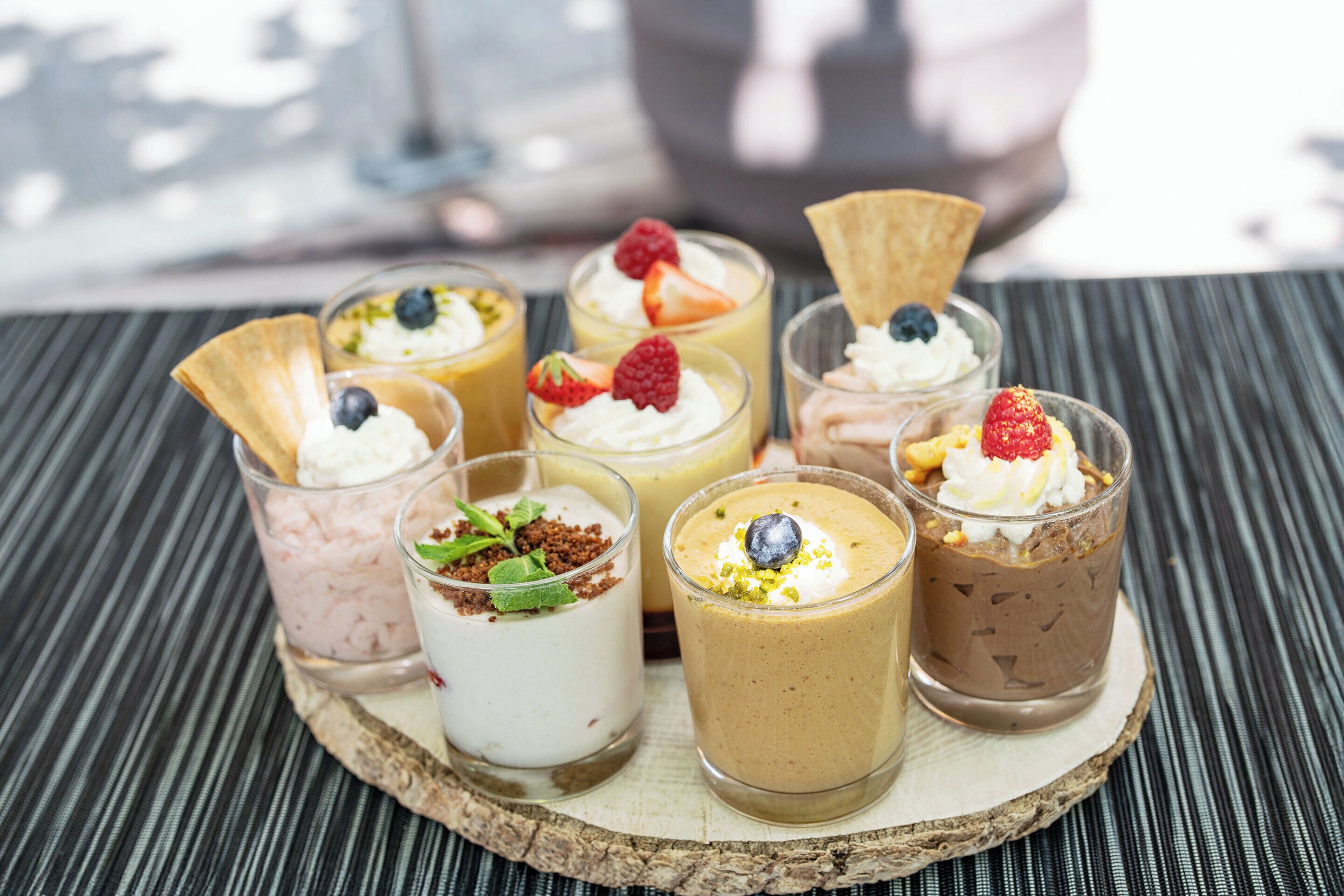 Een dienblad met zes toetjes en diverse toppings, op een houten plank.