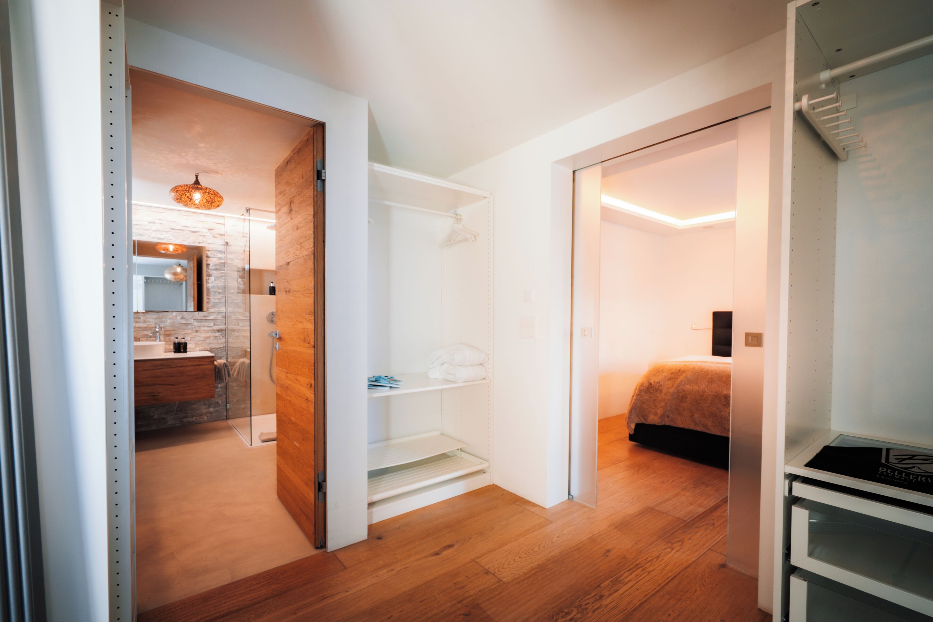 A modern hotel room with a wooden floor, a glass shower enclosure, a bed with a white headboard, and a small kitchenette area.