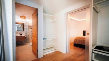 A modern hotel room with a wooden floor, a glass shower enclosure, a bed with a white headboard, and a small kitchenette area.