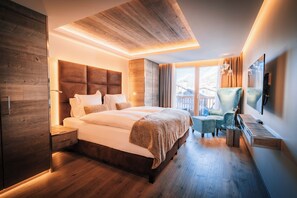 A modern hotel room with a large bed, wooden flooring, a seating area with a chair and table, and a large window offering a view. - Hotel Bellerive (Zermatt)