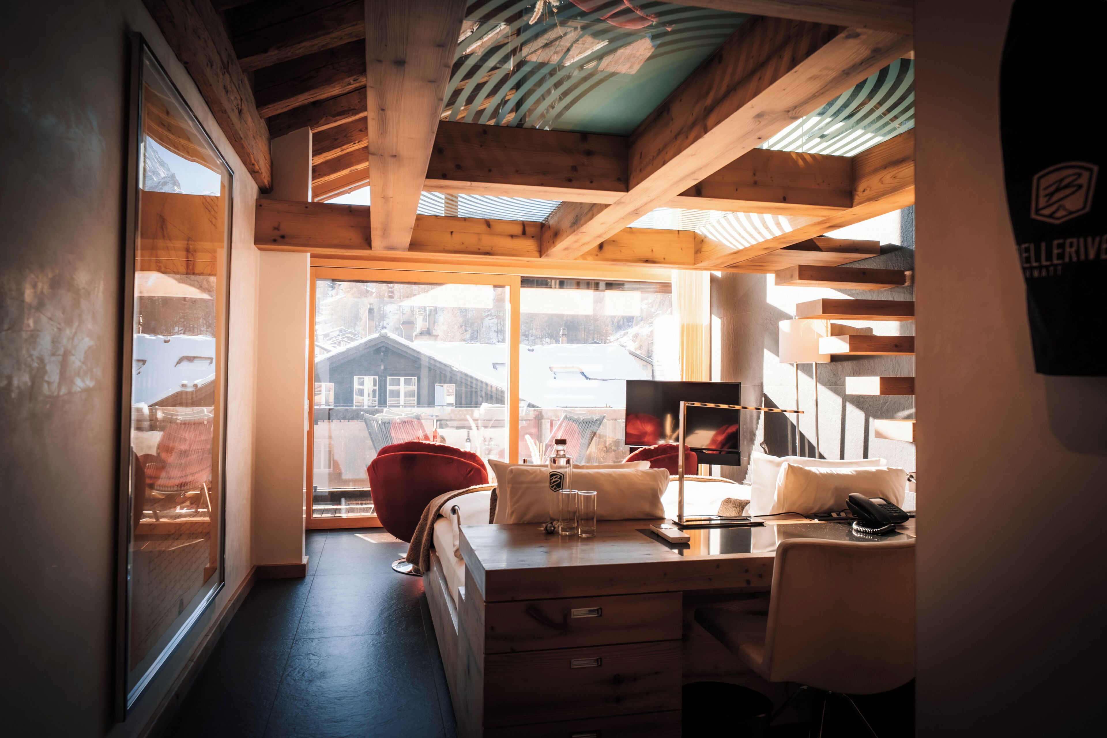 A room with a wooden desk, a chair, a bed, and a large window with a view of buildings outside.