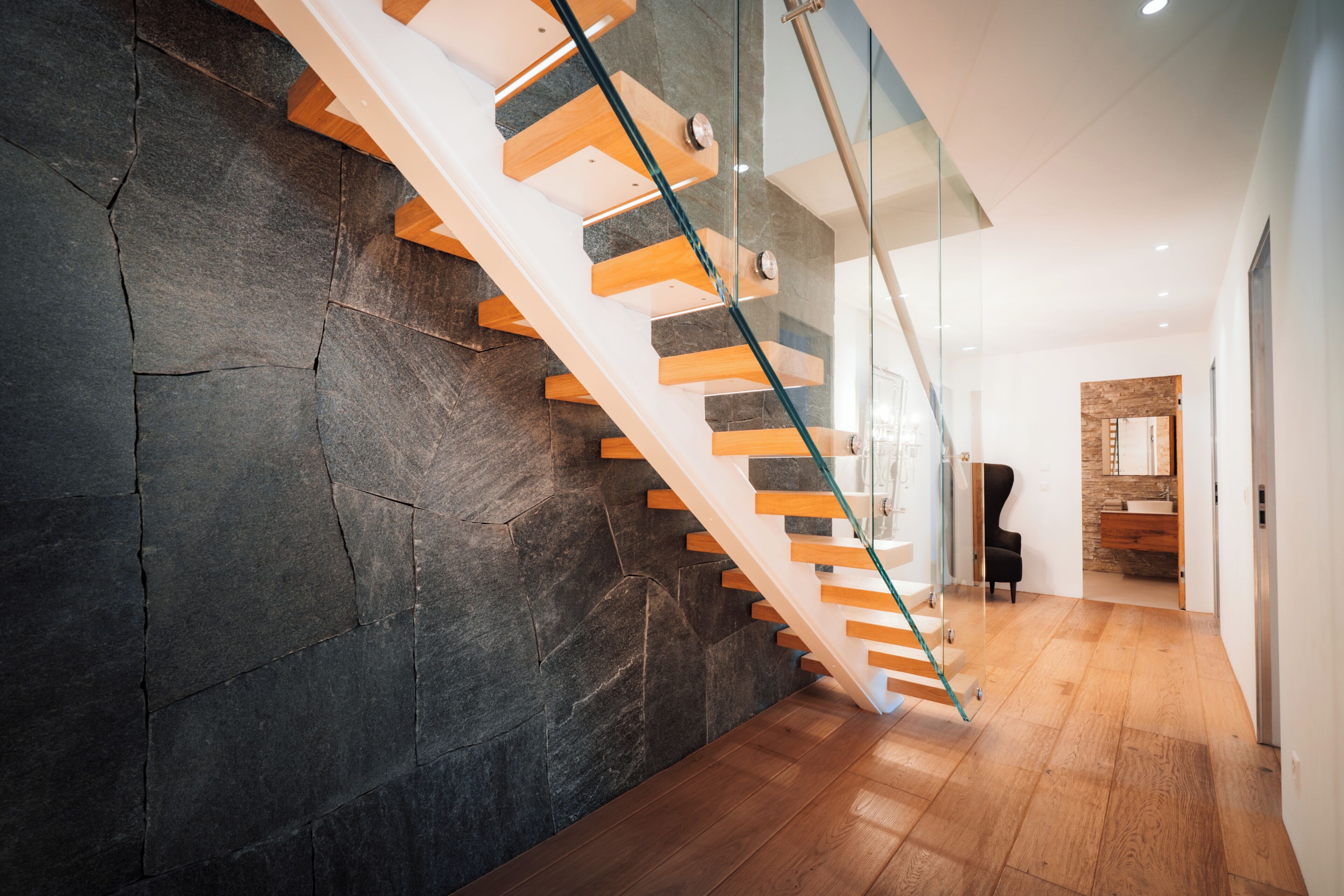Una escalera moderna con barandilla de vidrio, peldaños de madera y pared de piedra.