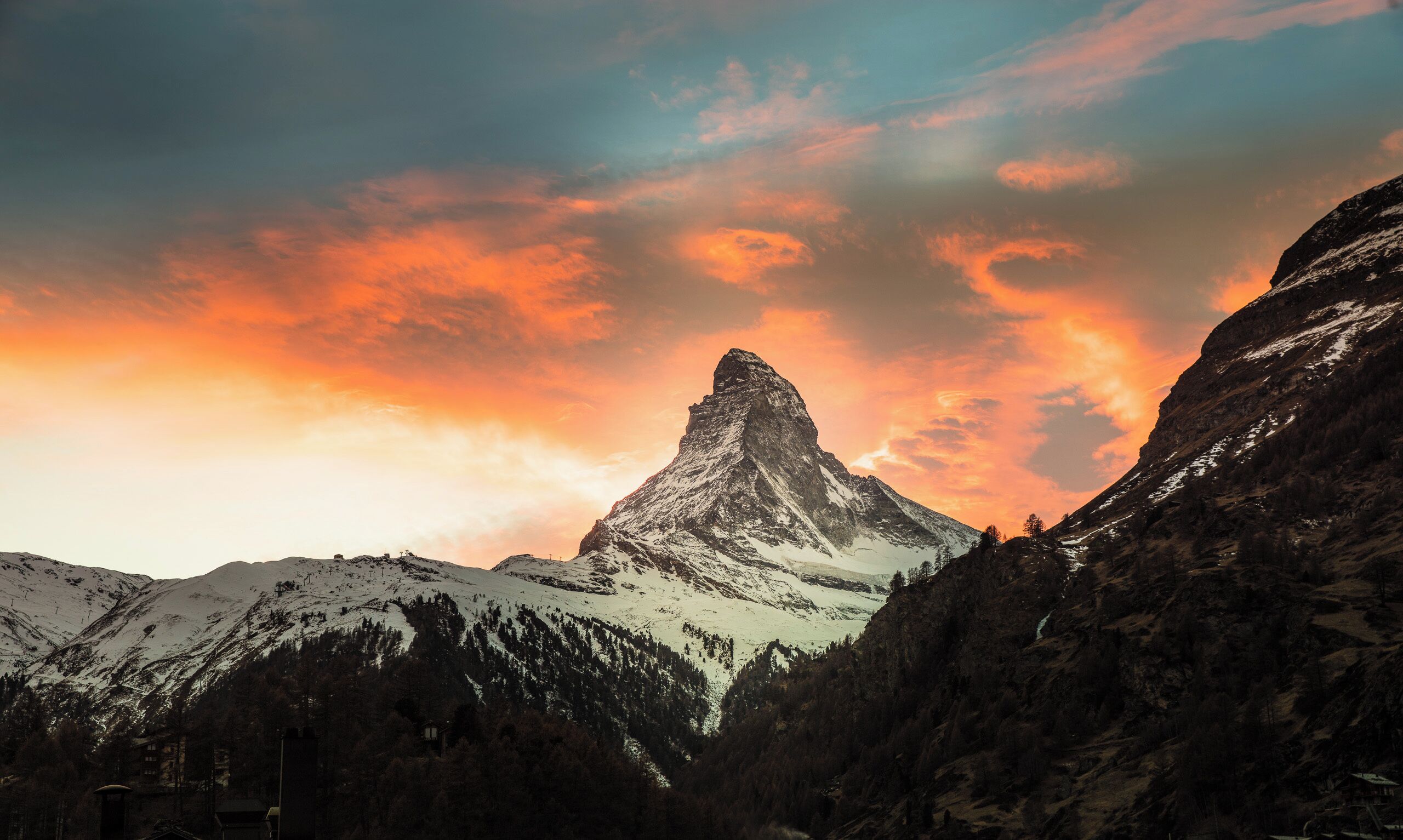 A mountain peak with a snow-covered summit, surrounded by forested slopes and a clear sky.