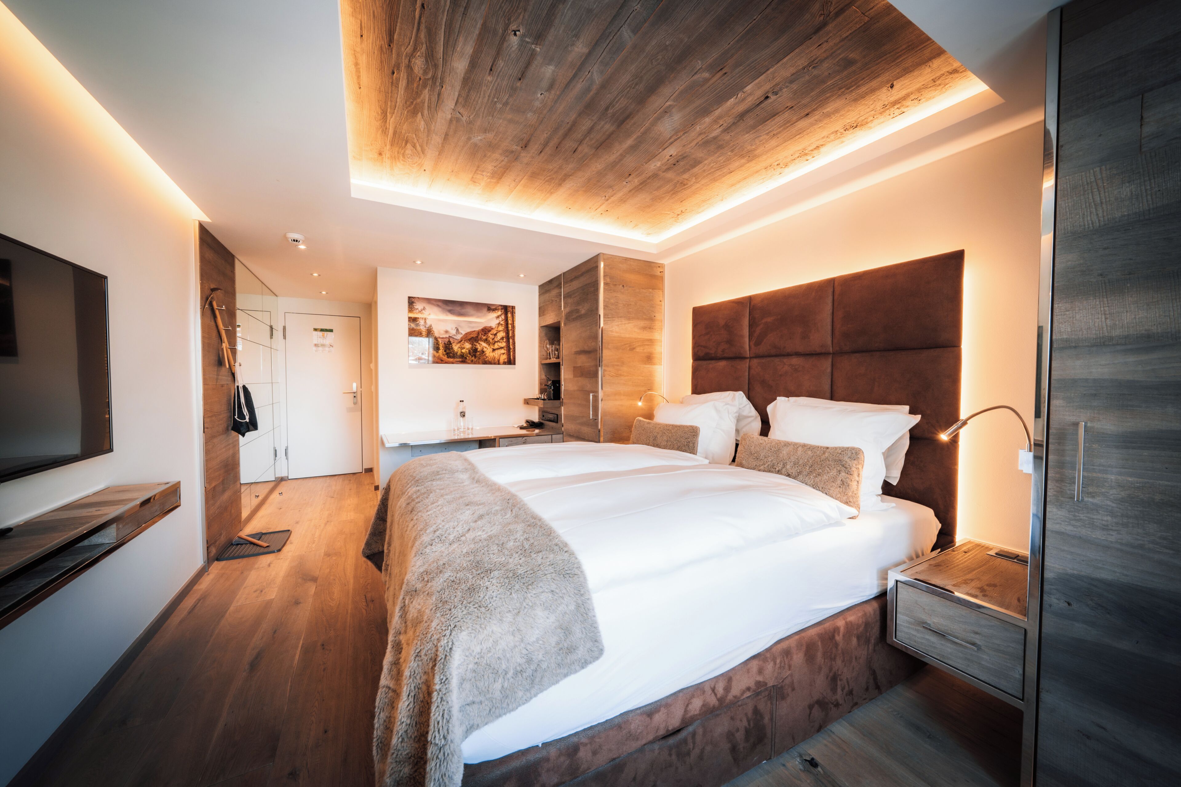 A modern bedroom with a large bed, wooden ceiling, and a mounted TV.