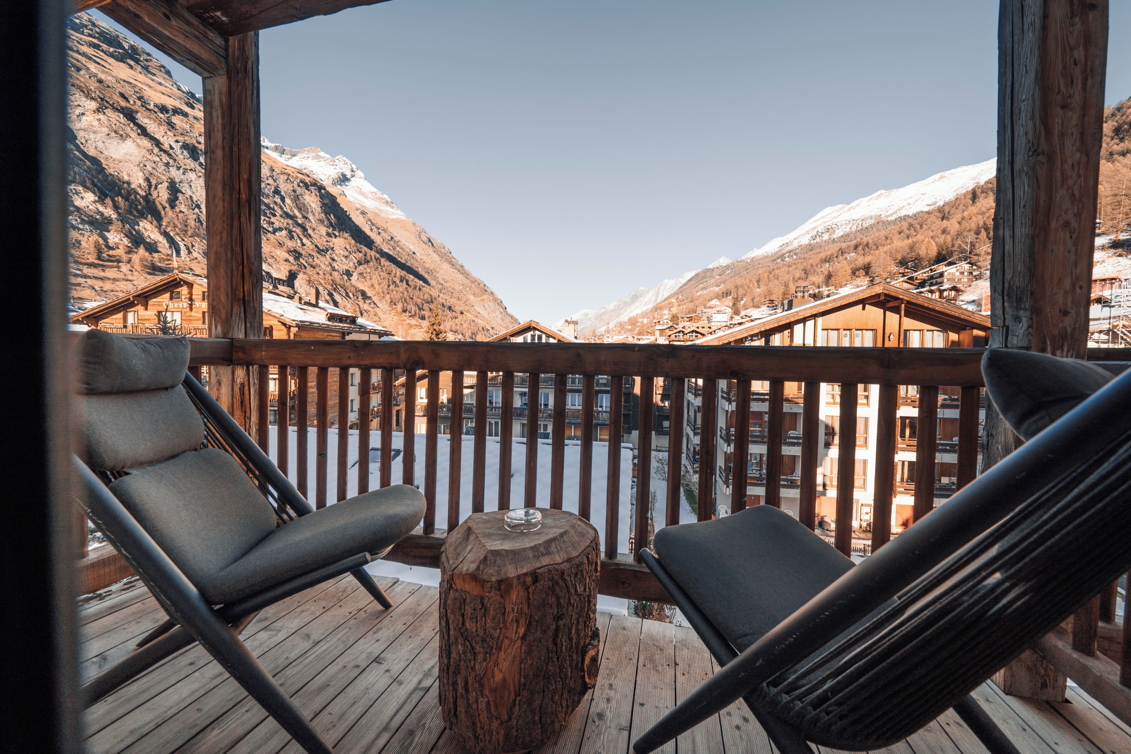 Eine Holzterrasse mit zwei Liegestühlen und einem Holztisch, mit Blick auf eine verschneite Berglandschaft und ein Dorf.