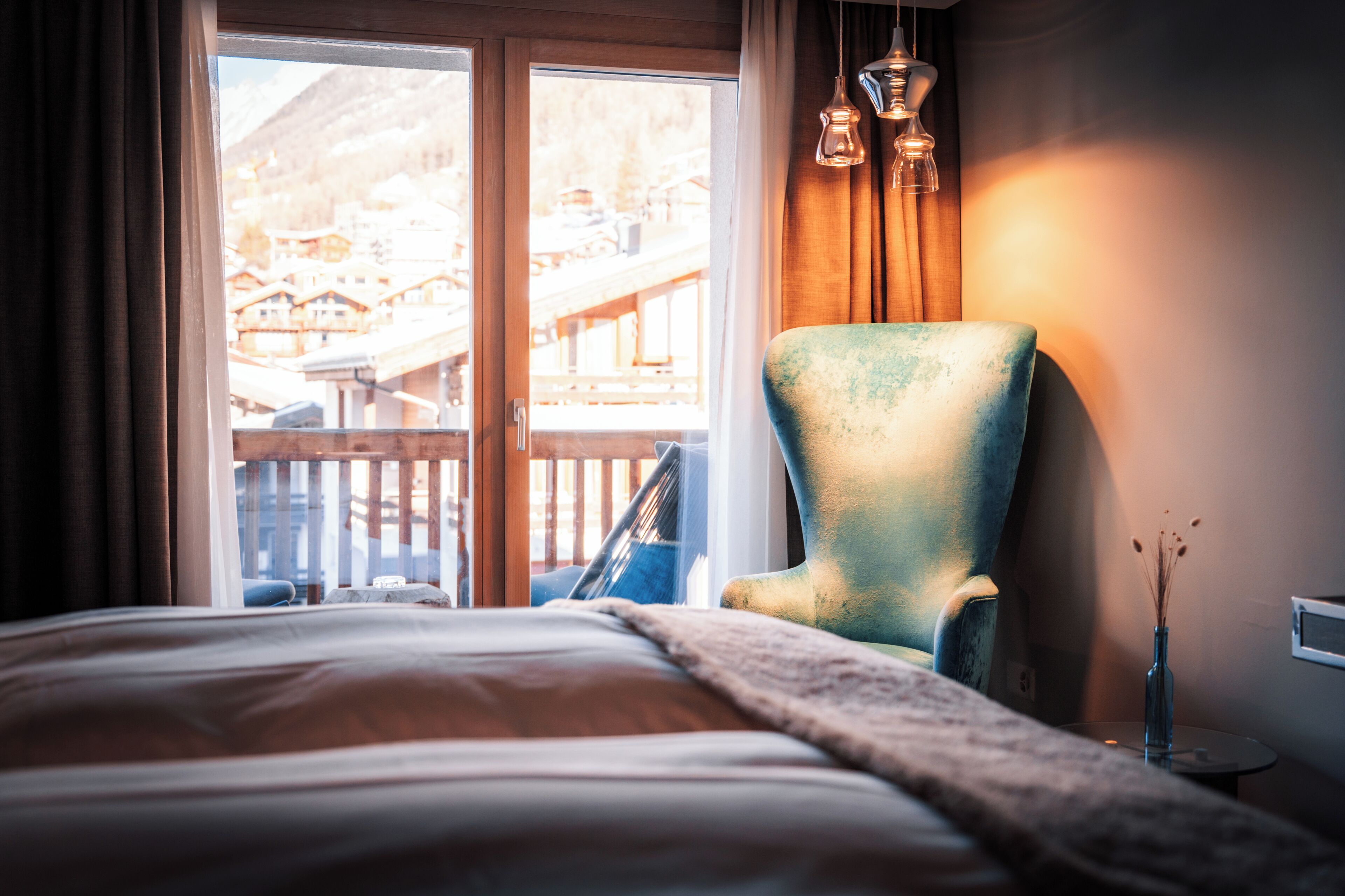 A hotel room with a bed, a chair, a vase with dried flowers, and a view of a building through a window.
