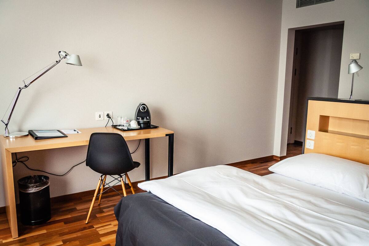 A hotel room with a wooden desk, a black office chair, a bed with white bedding, and a nightstand with a lamp.