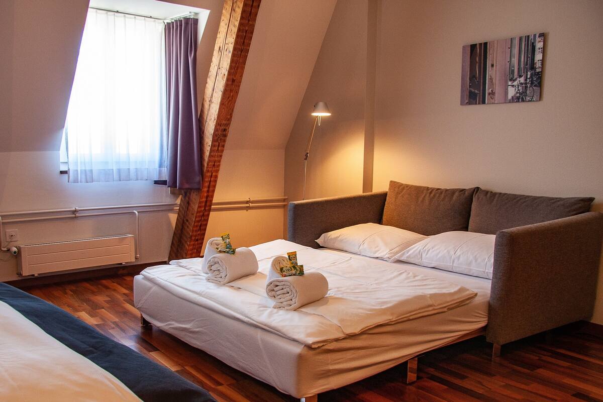 A bedroom with a bed, a bedside table, a lamp, a window with curtains, and a painting on the wall.