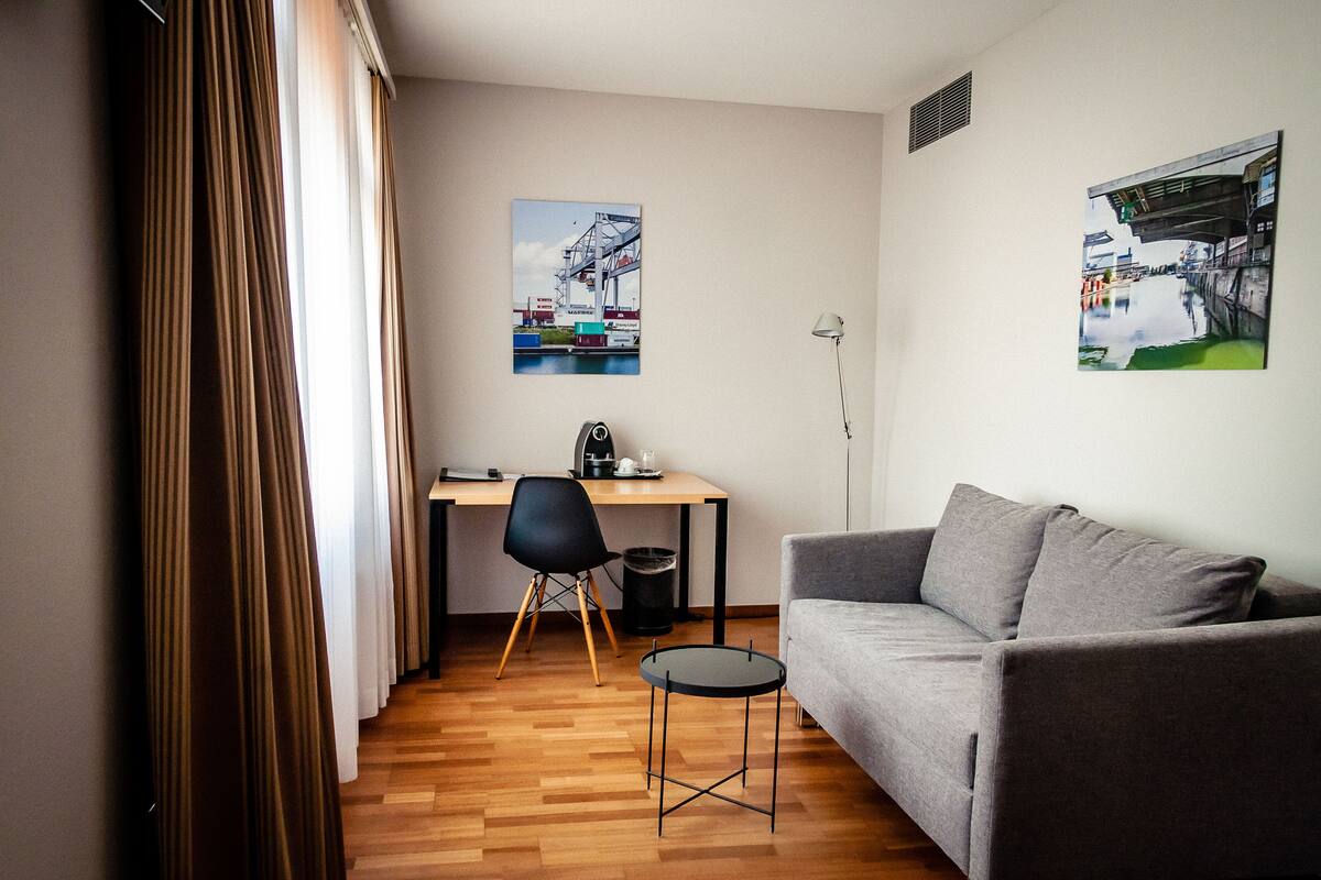 A modern living room with a grey sofa, a small round table, a black chair, and a desk with a computer. There are two wall-mounted pictures depicting urban scenes.