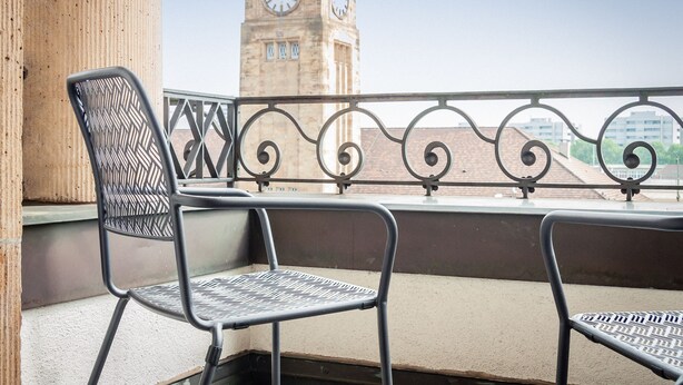 A balcony with two metal chairs and a wrought iron railing, overlooking a clock tower. - Royal Hotel (Basel)