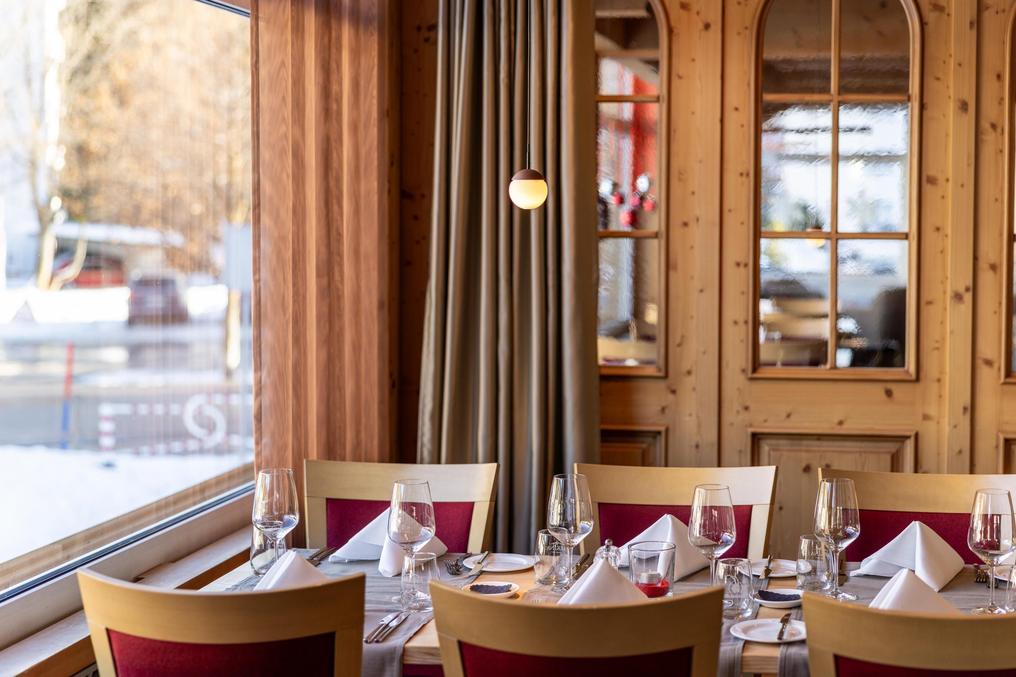 Un restaurante con paneles de madera, vistas a la calle por una ventana, y mesas preparadas con copas de vino, platos y cubiertos.