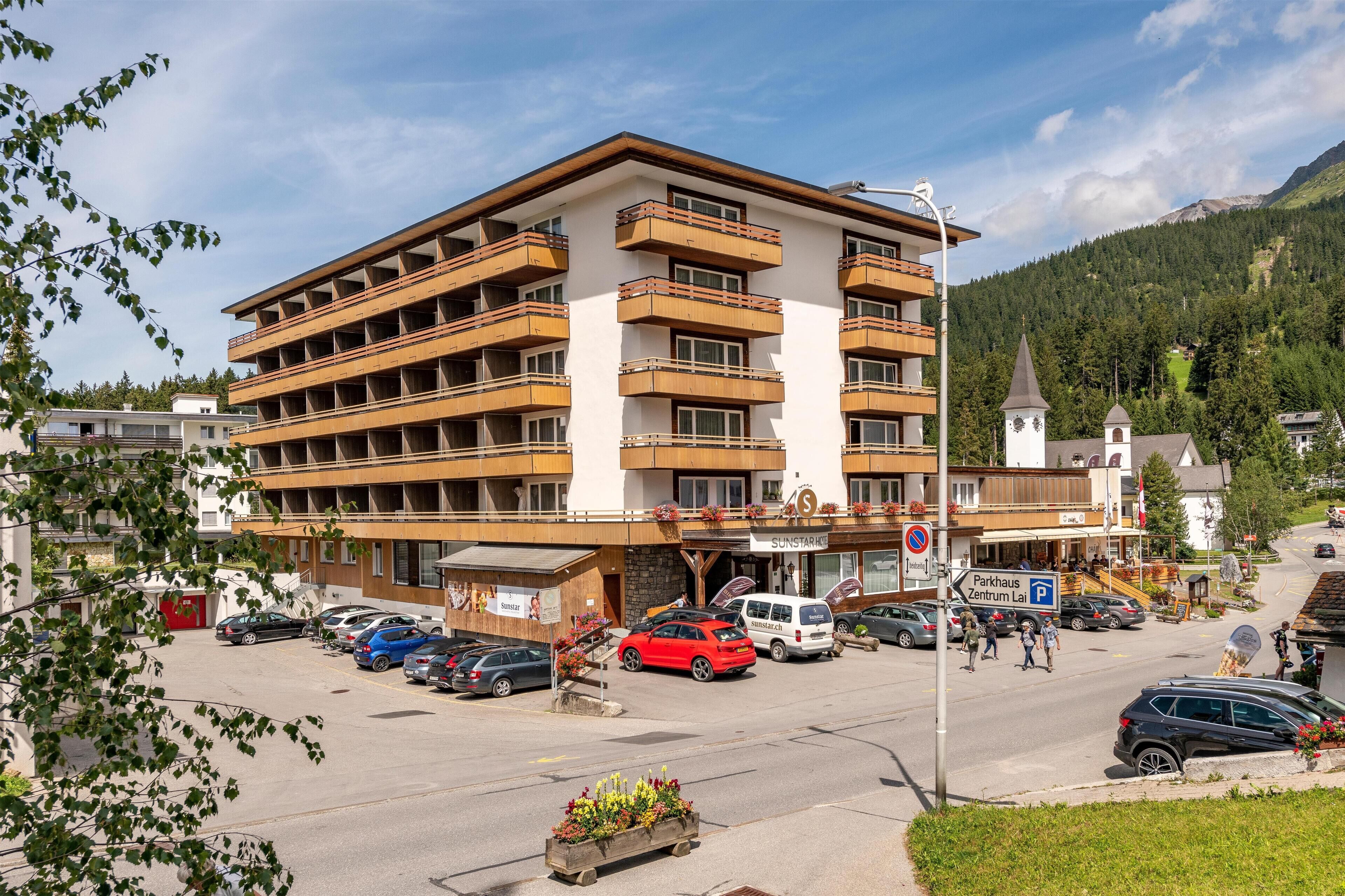A multi-story hotel with balconies, a parking area, and a view of a mountain.