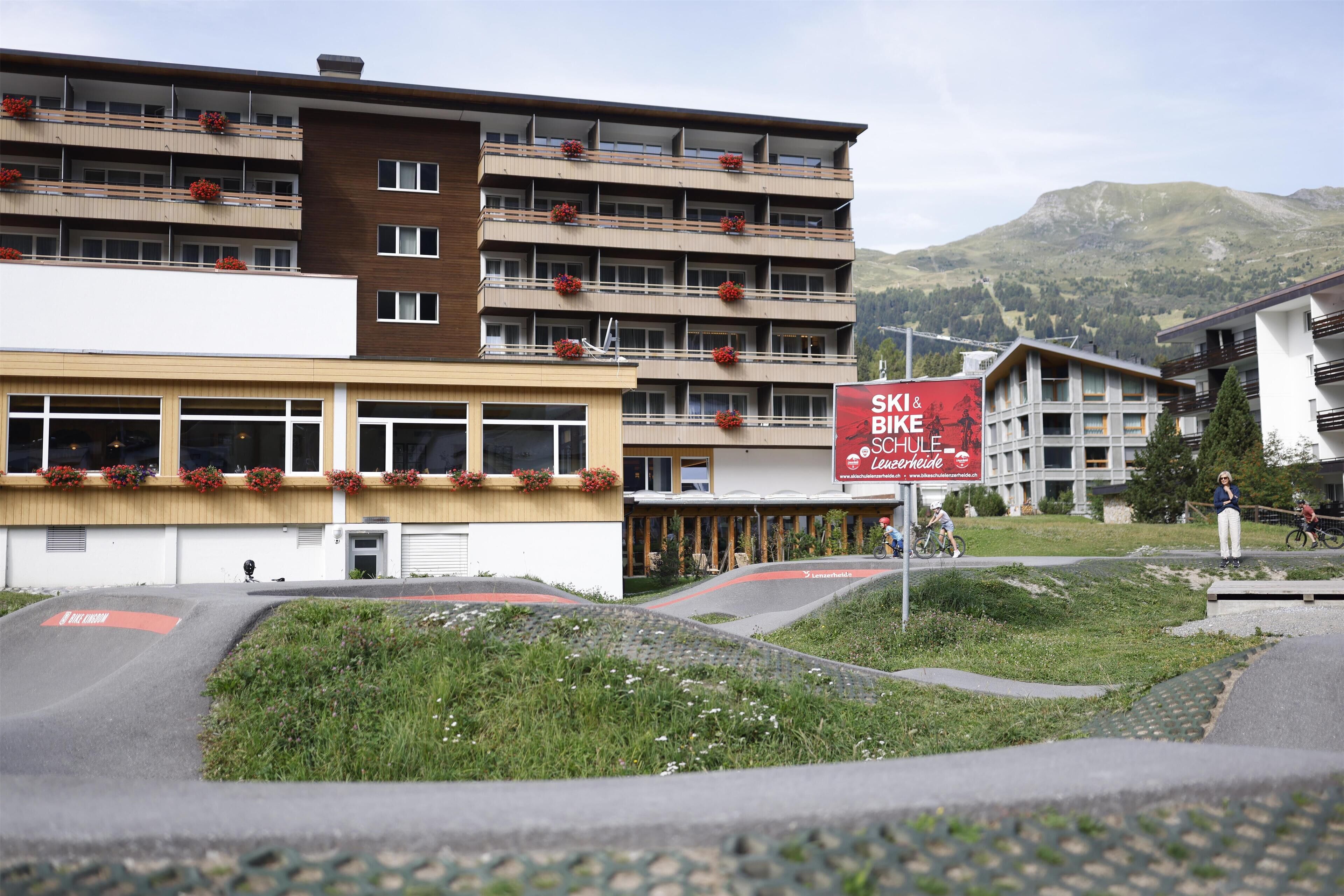 Ein mehrstöckiges Hotel mit Balkonen, die mit roten Blumen geschmückt sind, ein Schild für Skier- und Fahrradverleih und eine gebirgige Landschaft im Hintergrund.