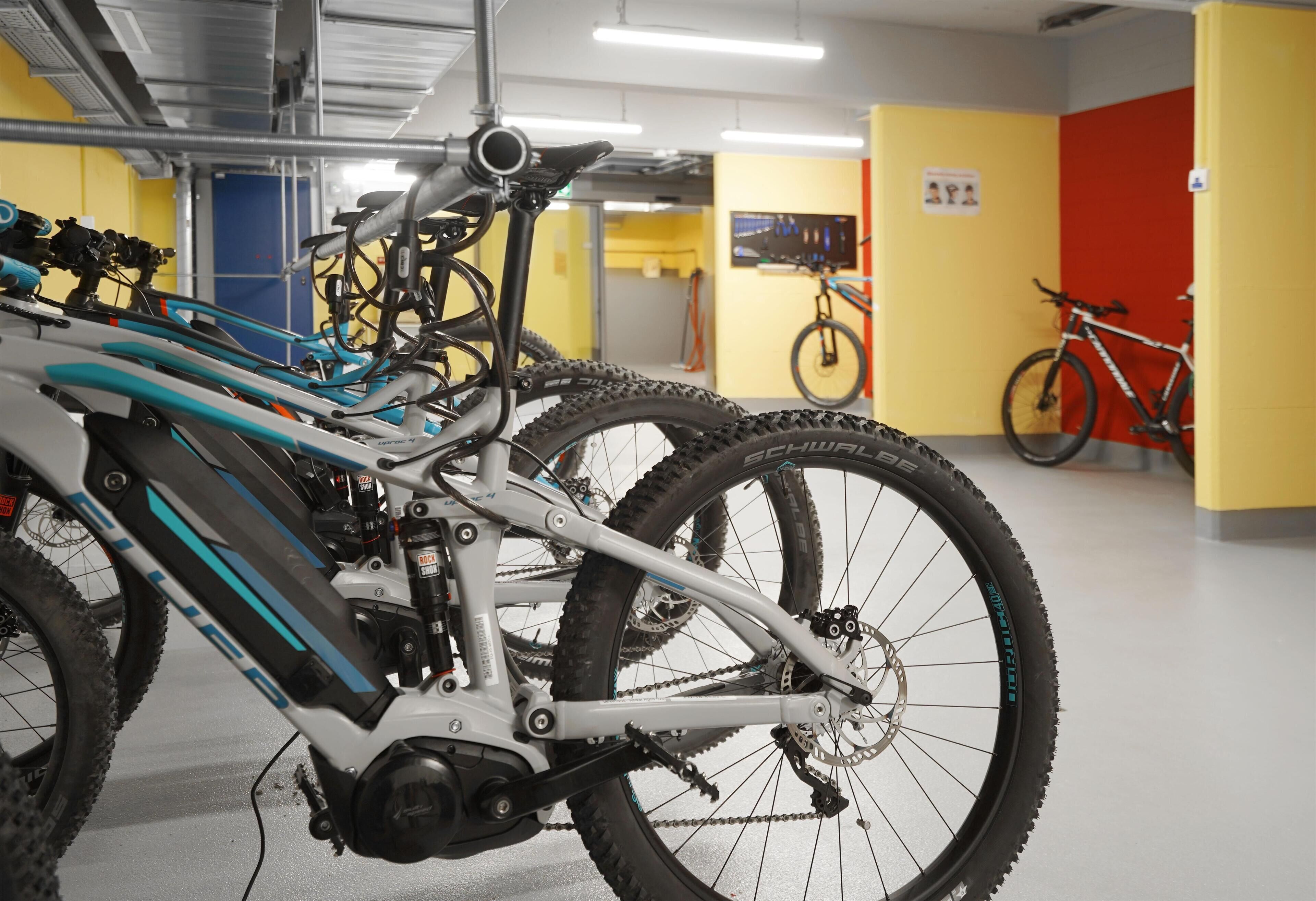 Una estación de reparación de bicicletas con herramientas y una bicicleta en primer plano, además de otras bicicletas y un televisor al fondo.