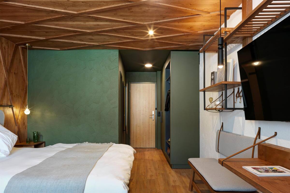 A bedroom with a wooden ceiling, a bed with a grey blanket, a wooden bench with a magazine, and a TV mounted on the wall.