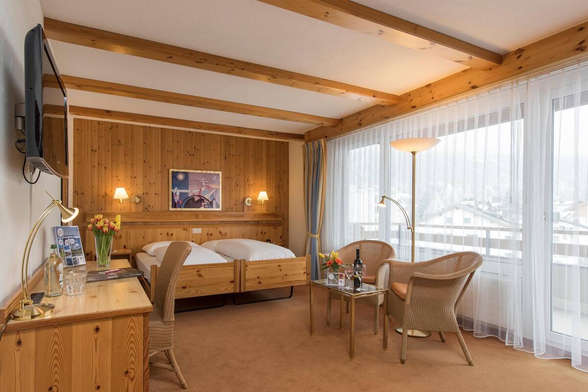 A hotel room with wooden panel walls, a bed, a desk with a lamp, a chair, and a large window with sheer curtains.