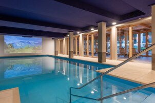 Una piscina interior con paredes de vidrio transparente, vigas de madera y una proyección de un paisaje montañoso.