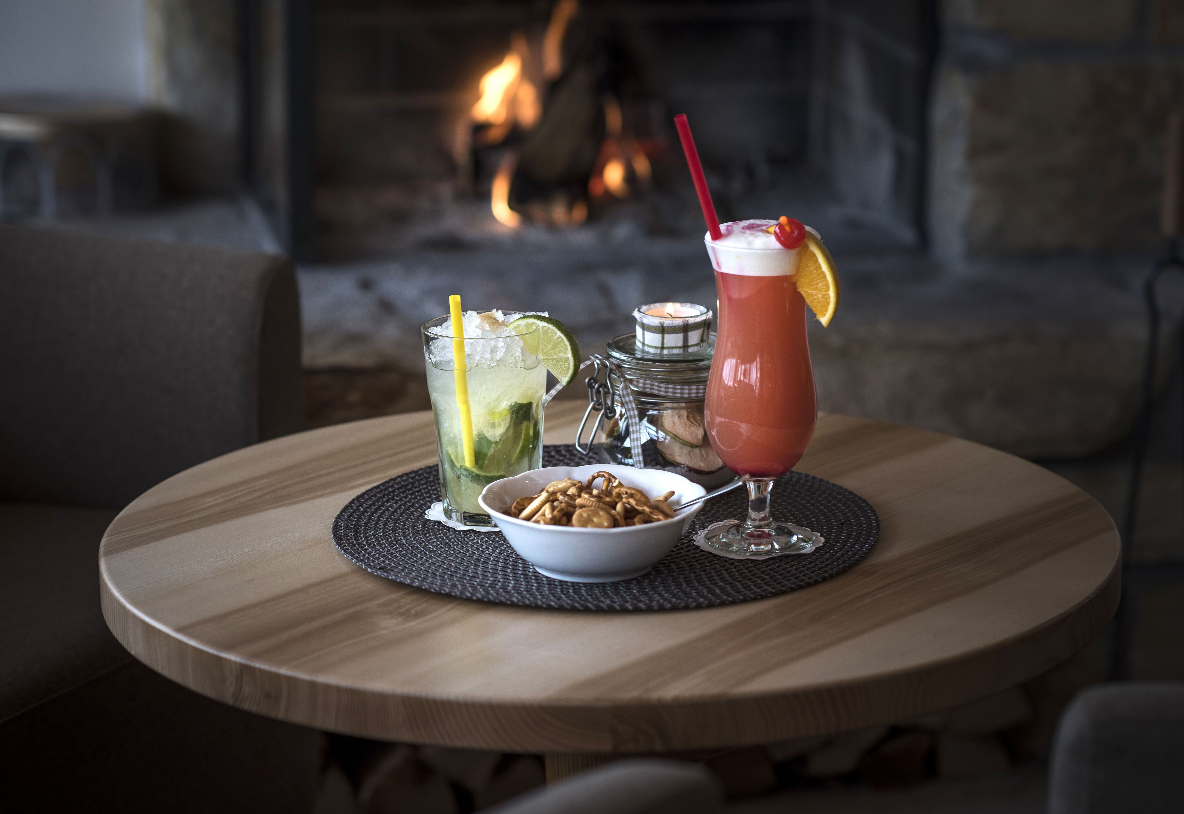 Una mesa redonda de madera con un bol de frutos secos, dos bebidas y una vela.
