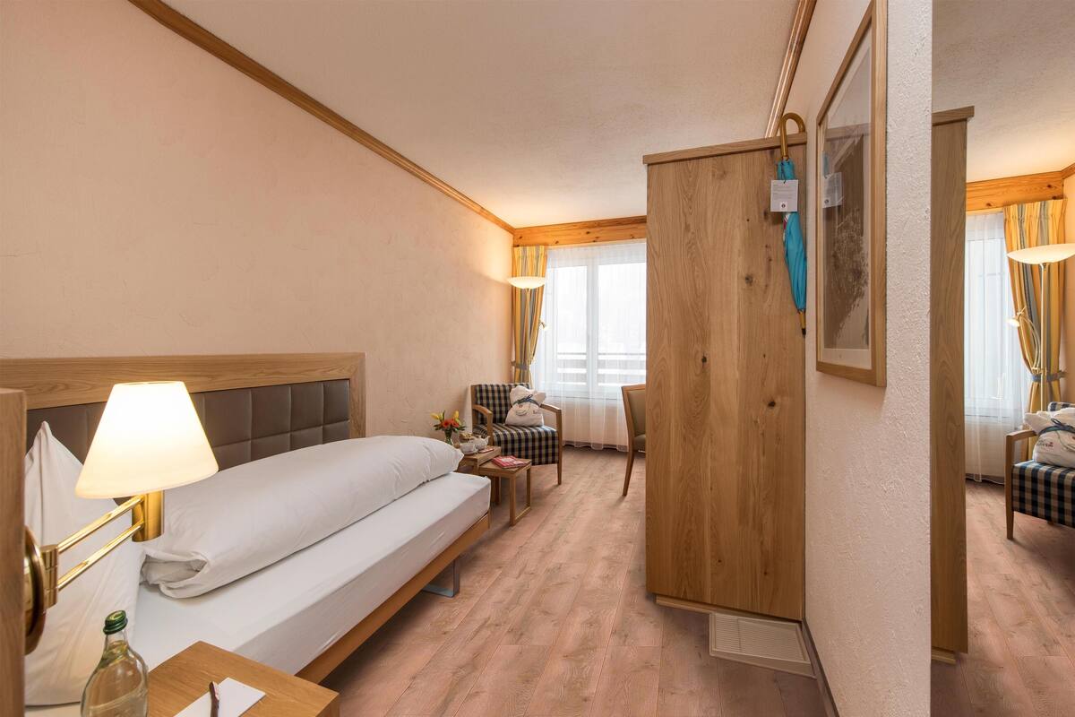 A hotel room with a bed, bedside lamp, wooden wardrobe, armchair, and a small table with a bottle and a notepad.