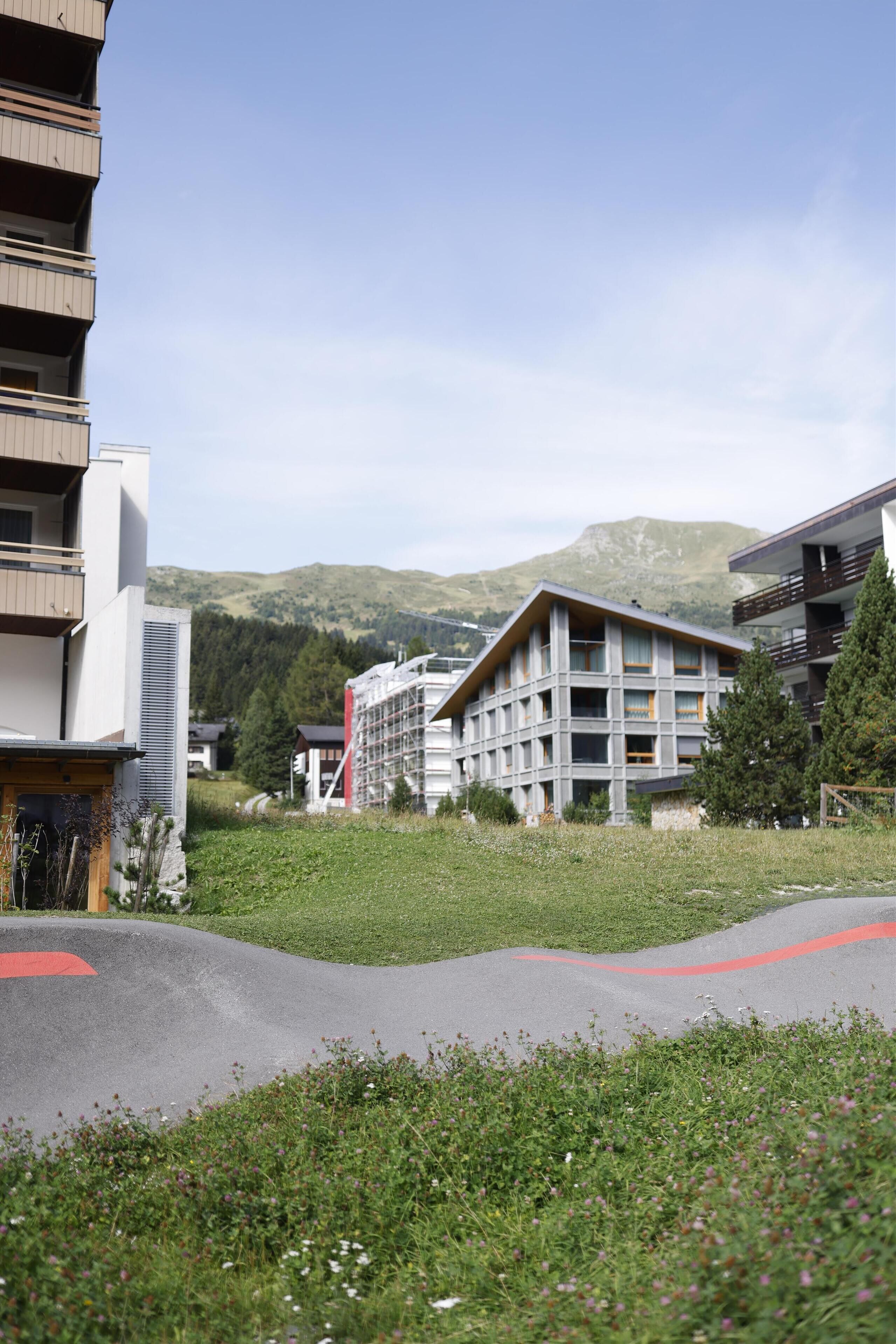 Un bâtiment moderne doté de nombreux balcons, un espace verdoyant avec un chemin rouge et un paysage montagneux en arrière-plan.