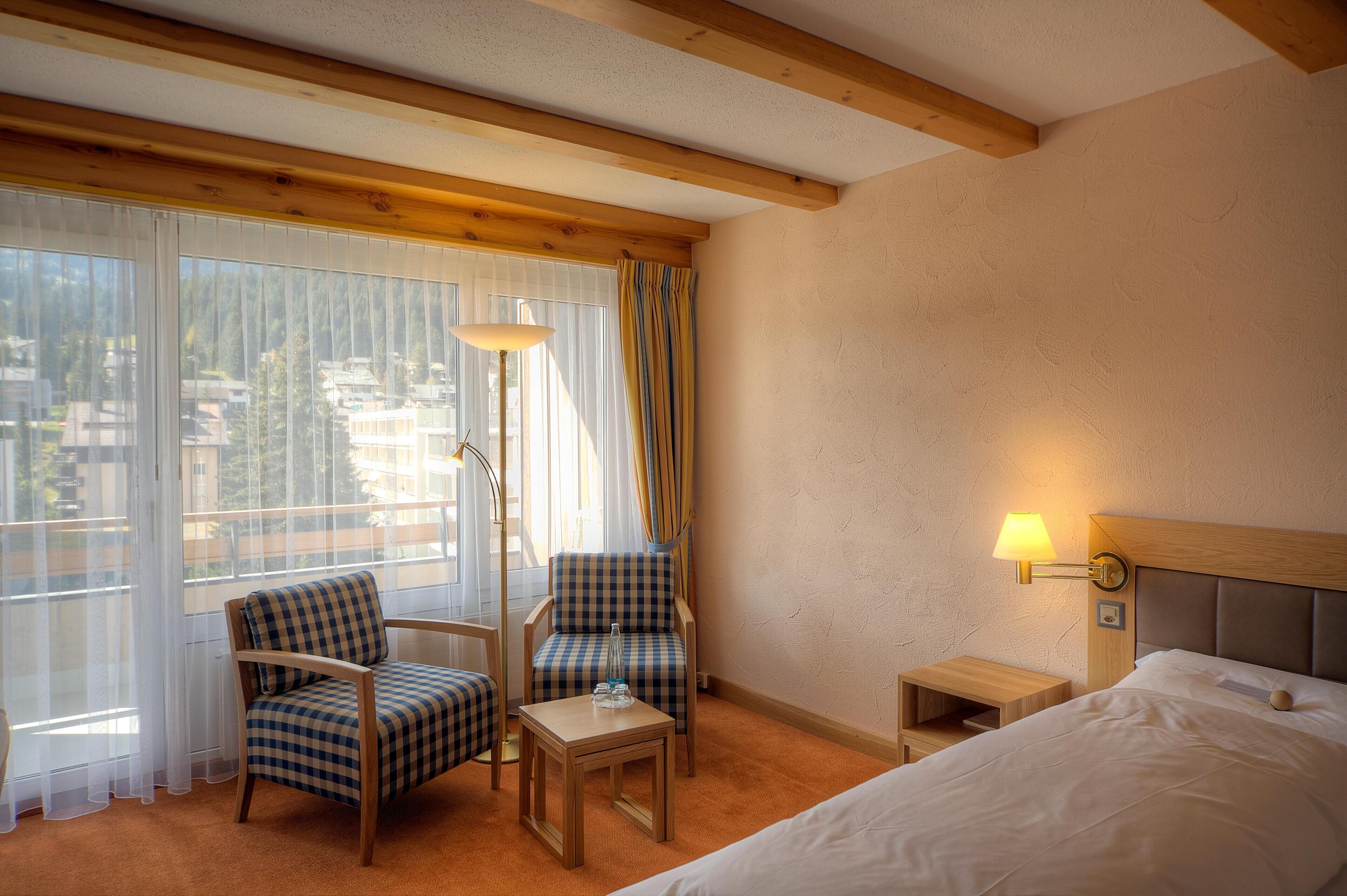 Une chambre d’hôtel avec un lit, deux fauteuils à motifs à carreaux, une petite table, une table de chevet, une lampe et de grandes fenêtres avec des rideaux légers.