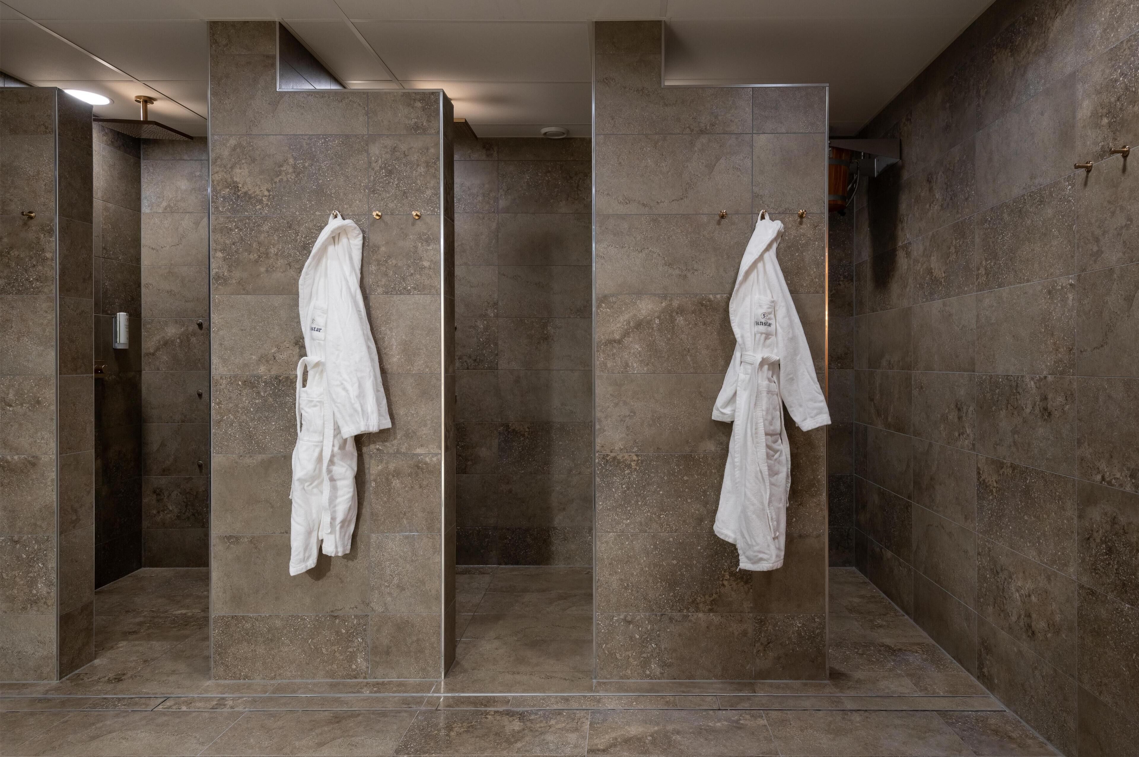 A modern bathroom with large stone tiles, a glass shower enclosure, and two white robes hanging on the wall.