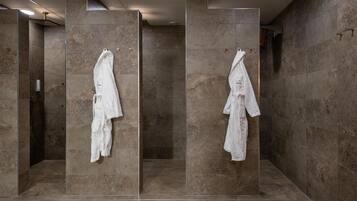 A modern bathroom with large stone tiles, a glass shower enclosure, and two white robes hanging on the wall.