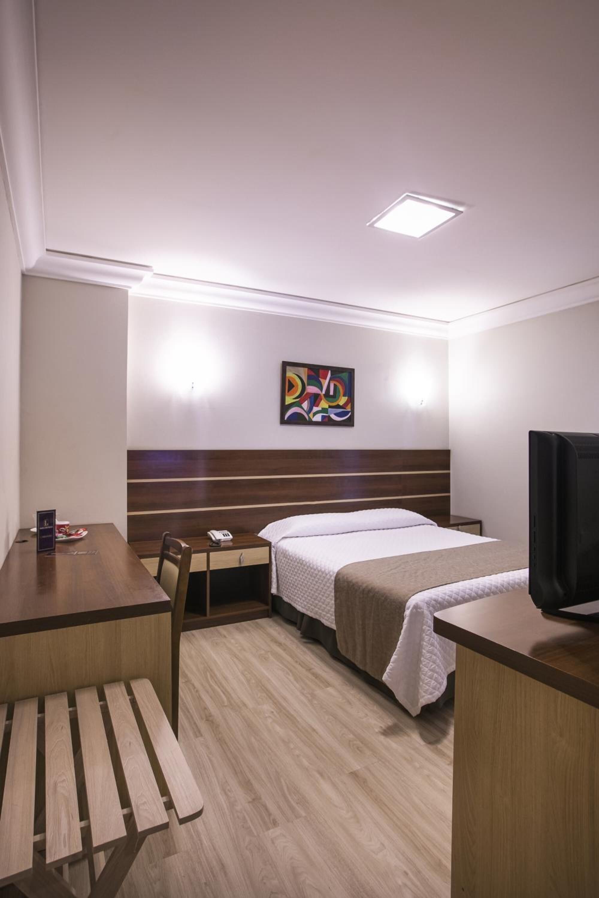 Quarto de hotel com cama, escrivaninha, cadeira, televisão e um quadro abstrato colorido na parede.