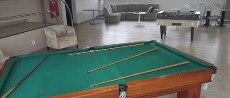 Una mesa de billar con tacos, un sofá y una mesa de fútbolín en una habitación con ventanas amplias.