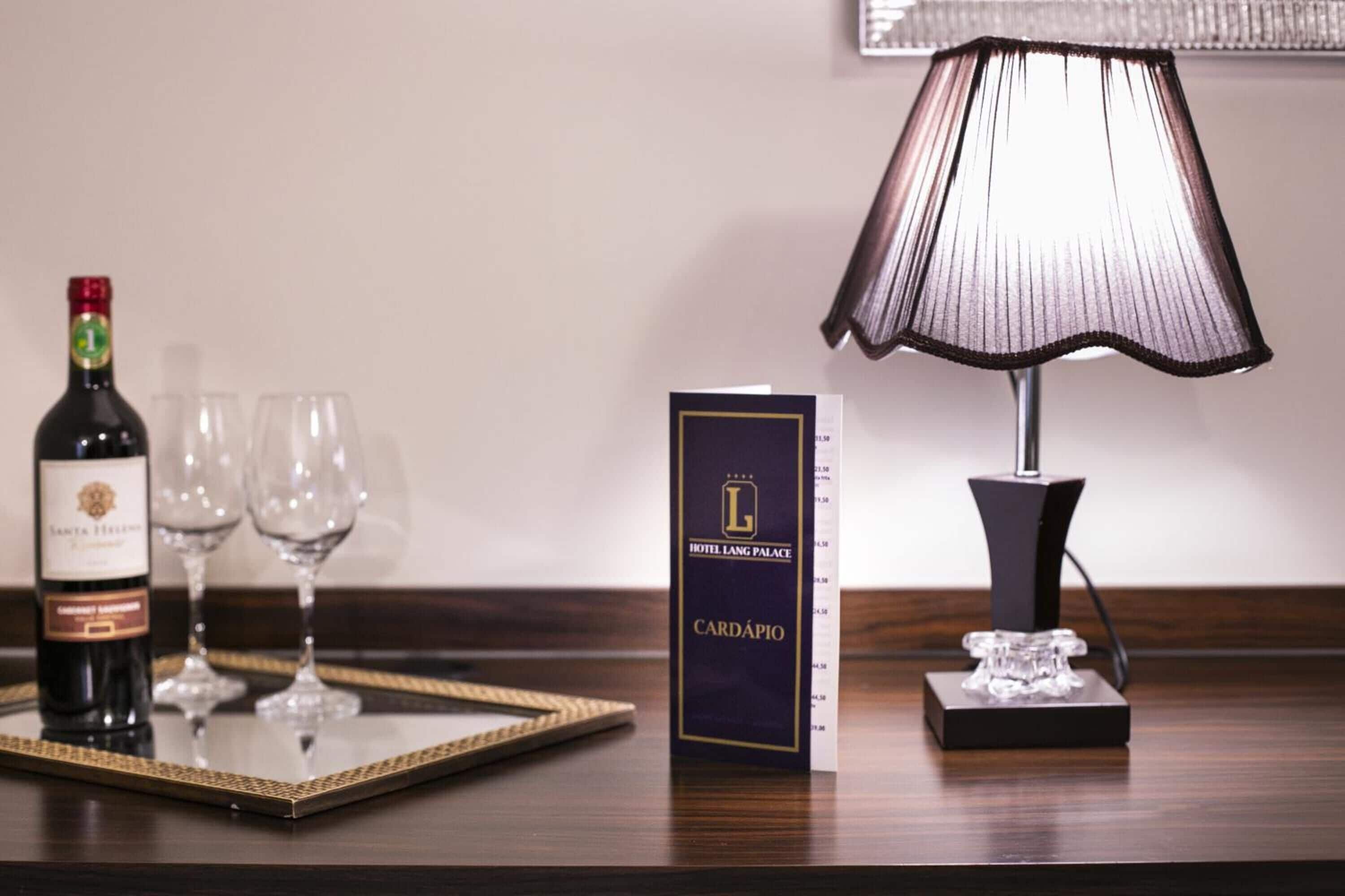 Habitación de hotel con una botella de vino, dos copas de vino, una lámpara y una tarjeta del Hotel Lang Palace.