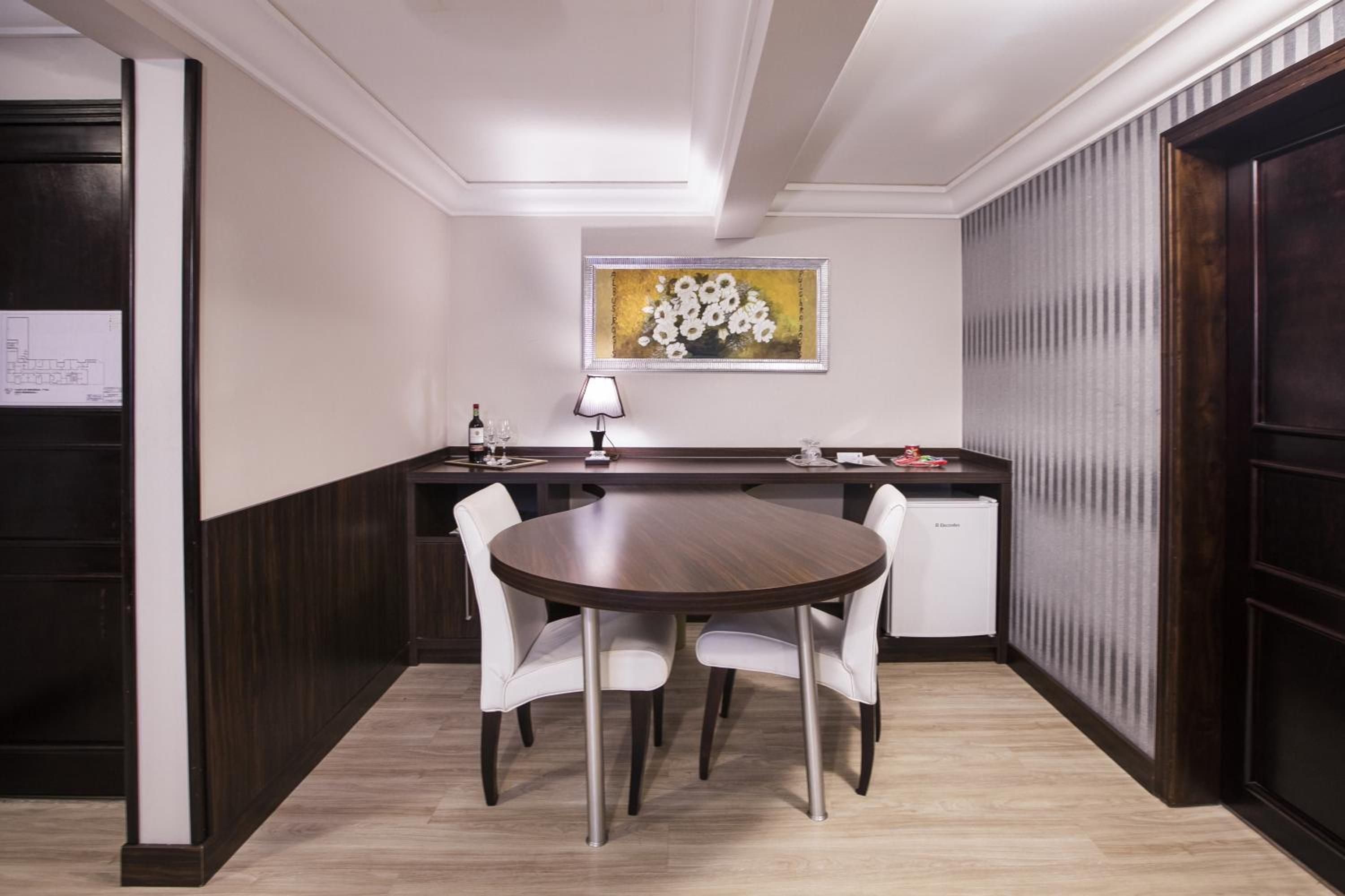 A dining area with a round table, two white chairs, a small table with a lamp, and a refrigerator.