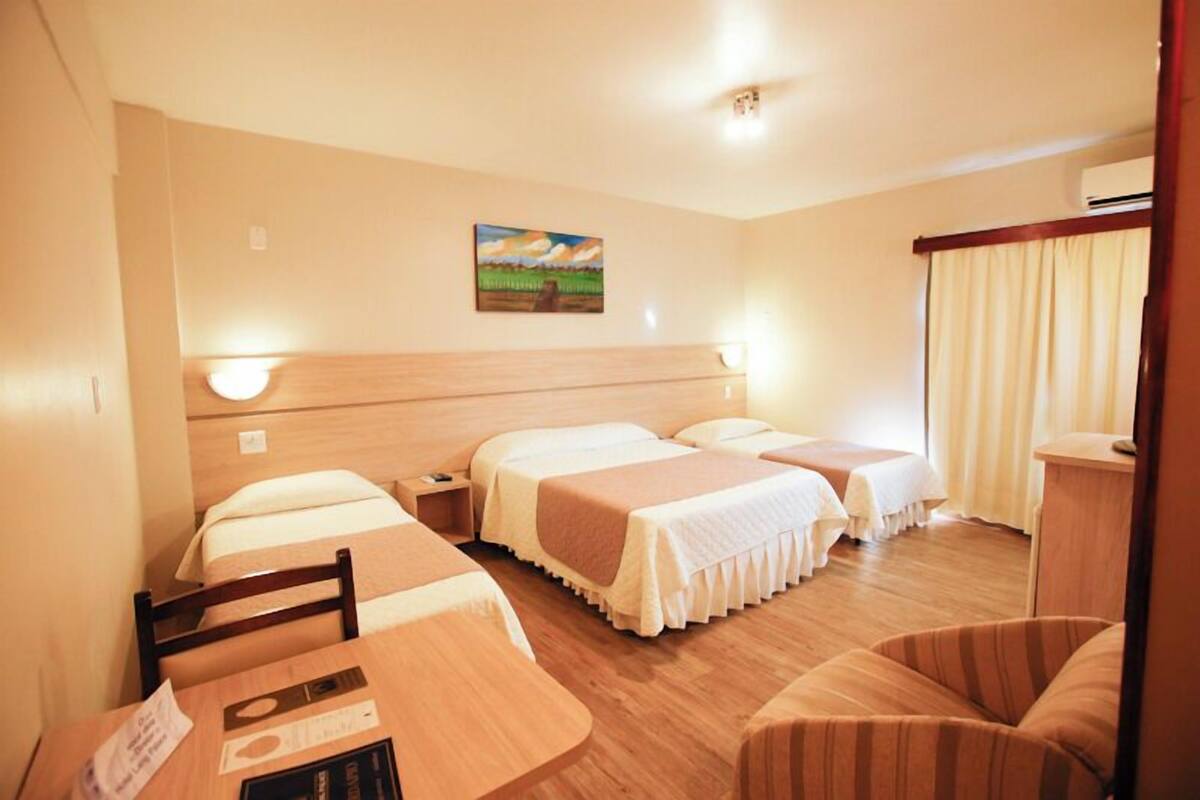 A hotel room with two beds, a desk, and a chair. There is a painting on the wall and a window with curtains.