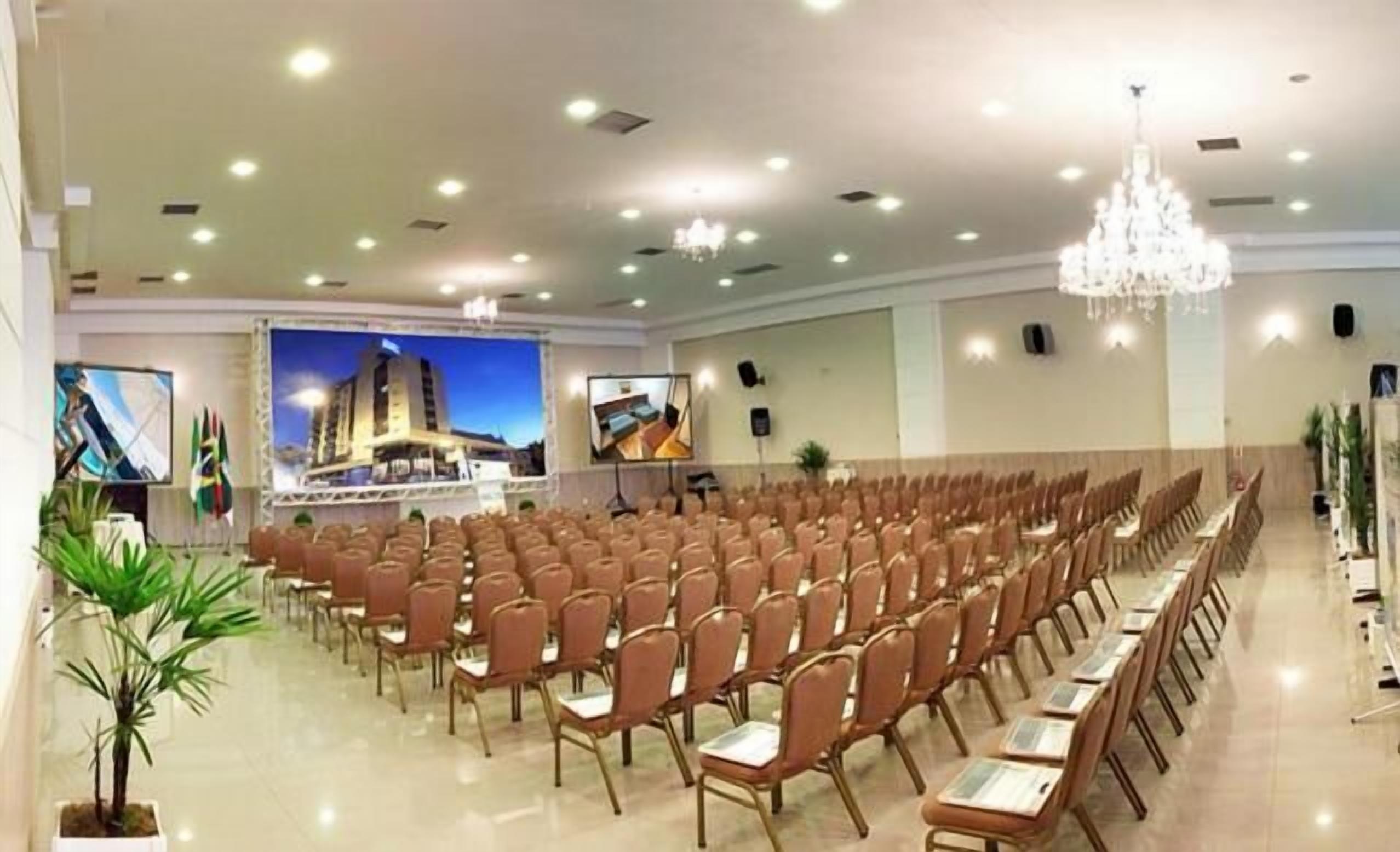 Un salón de conferencias con filas de sillas, un escenario con fondo y una araña.