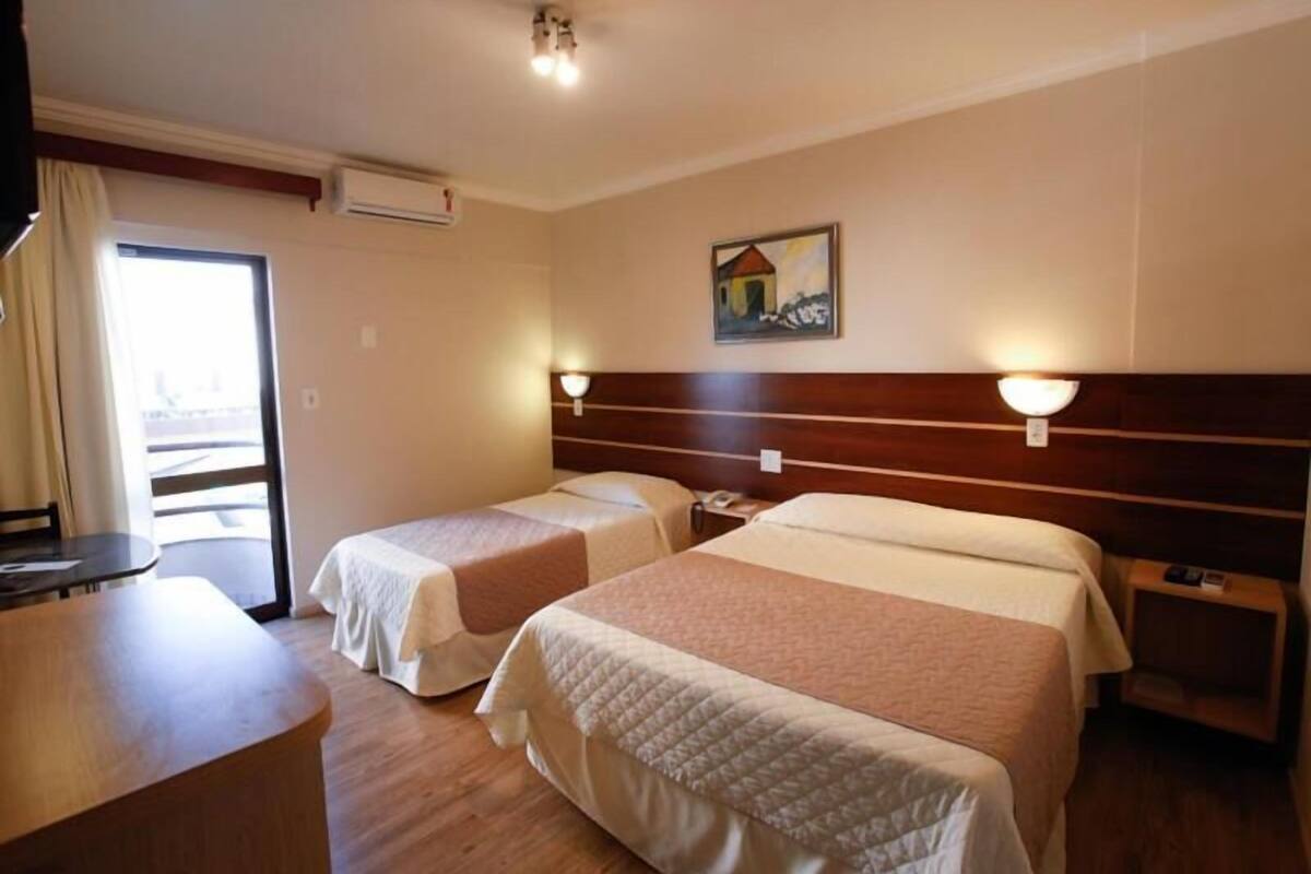 A hotel room with two beds, a wooden desk, a chair, a table, and a painting on the wall.