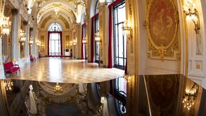 Un salón imponente con detalles dorados ornamentales, ventanas amplias y un suelo reflectante.