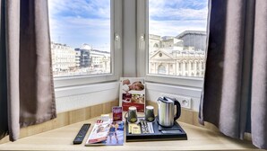 Vista desde la ventana de un paisaje urbano con edificios históricos, una mesa con control remoto, revistas, un cartel de Wi-Fi y una cafetera.