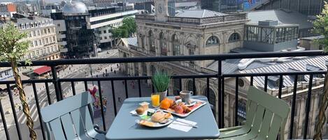 A balcony with a table set for breakfast, overlooking a cityscape with historic buildings and modern skyscrapers.