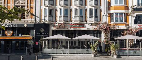 Un hôtel à la façade classique, doté de nombreux balcons et d’un café situé au rez-de-chaussée, avec une terrasse.