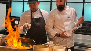 Dos cocineros en una cocina con un fuego grande en una sartén, equipo de cocina y una ventana con celosía.