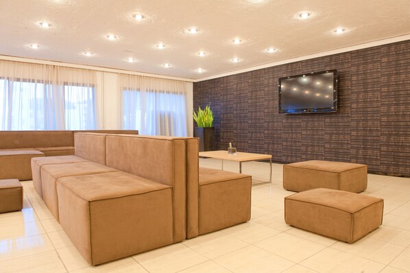 A modern living room with a sectional sofa, ottomans, a small table, and a flat-screen TV mounted on a textured wall. - Heronissos Hotel - All inclusive (Hersonissos)