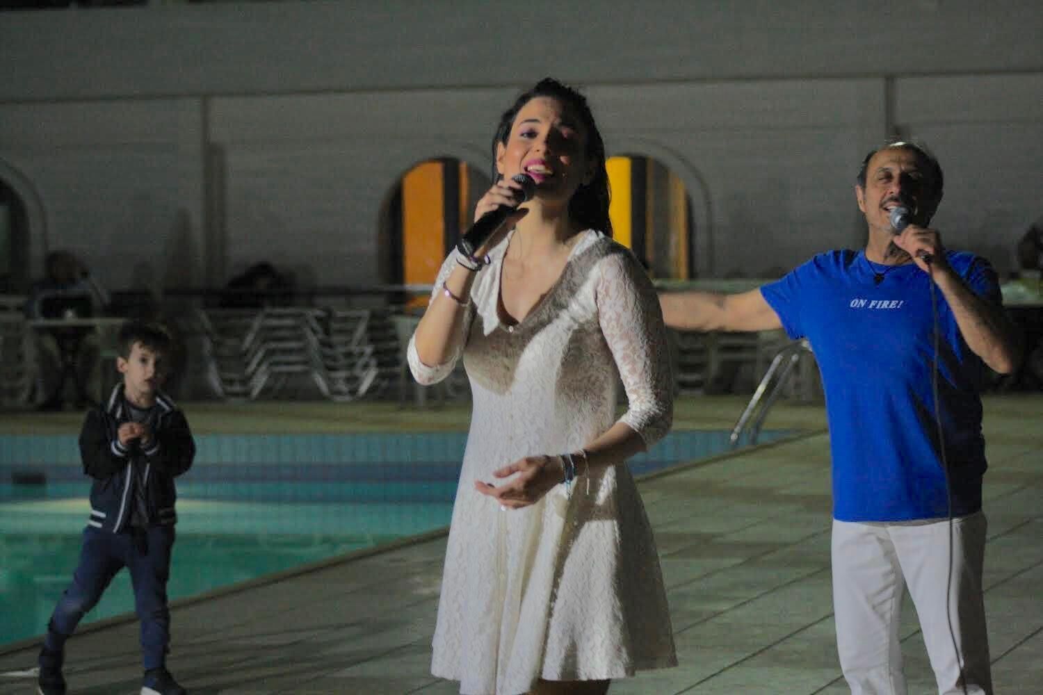 Une personne vêtue d’une robe blanche se tient près d’une piscine, une autre porte une chemise bleue, et un enfant est vêtu d’une veste noire.