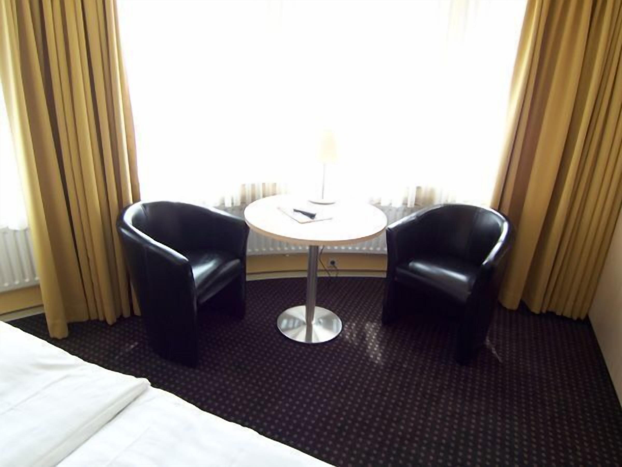 Habitación de hotel con una cama, dos sillones de cuero negro, una mesita redonda con una lámpara y una ventana con cortinas.