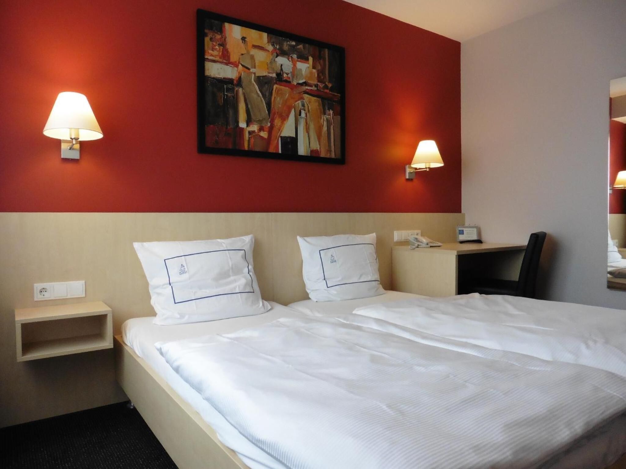 A hotel room with a double bed, two bedside tables, a desk with a chair, a painting on the wall, and two wall-mounted lamps.