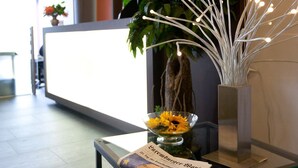 Ein Empfangsbereich mit Tischplatte aus Glas, einer Zeitung, einer Vase mit Sonnenblumen, einer Topfpflanze und einer dekorativen Lichtinstallation.