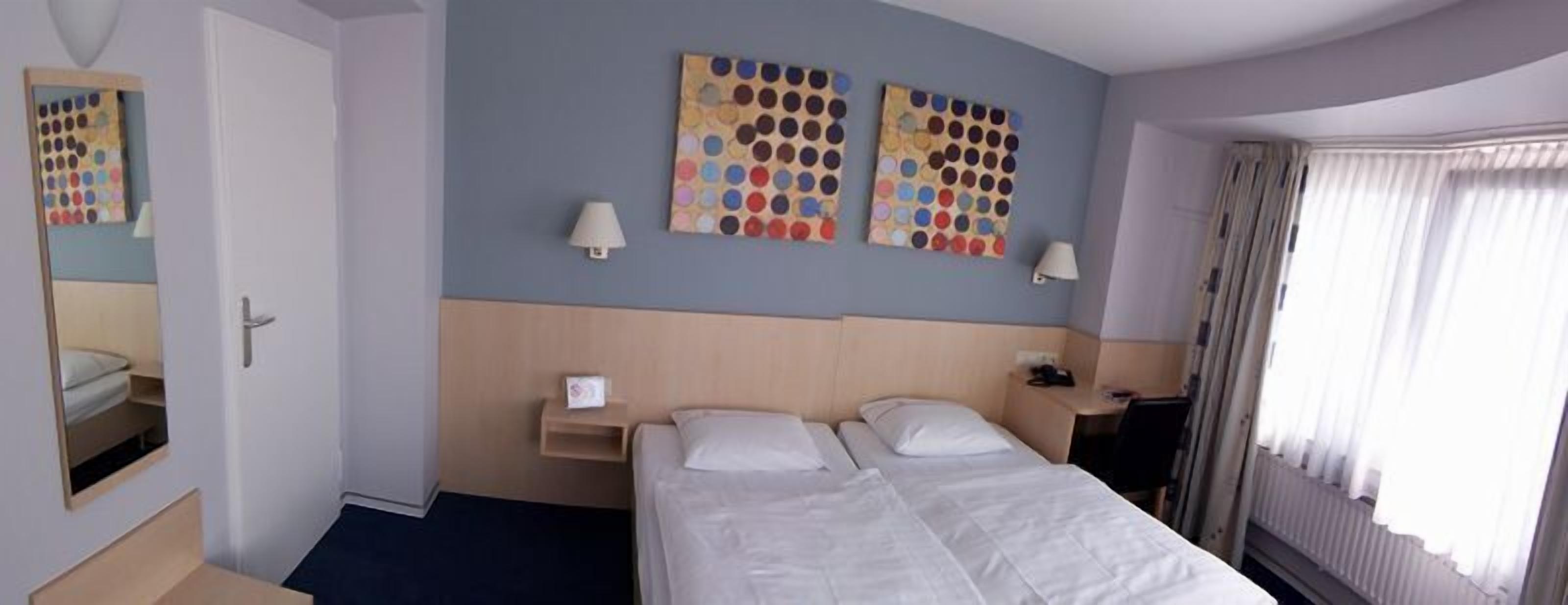 Una habitación de hotel con una cama doble, dos lámparas adosadas a la pared, una mesita de noche pequeña y un espejo.