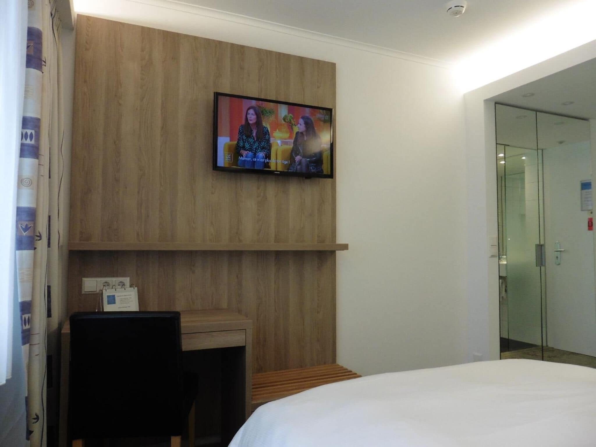 Ein Hotelzimmer mit einem Bett, einem Schreibtisch mit Stuhl, einem an der Wand befestigten Fernseher und einer Glastür zum Badezimmer.