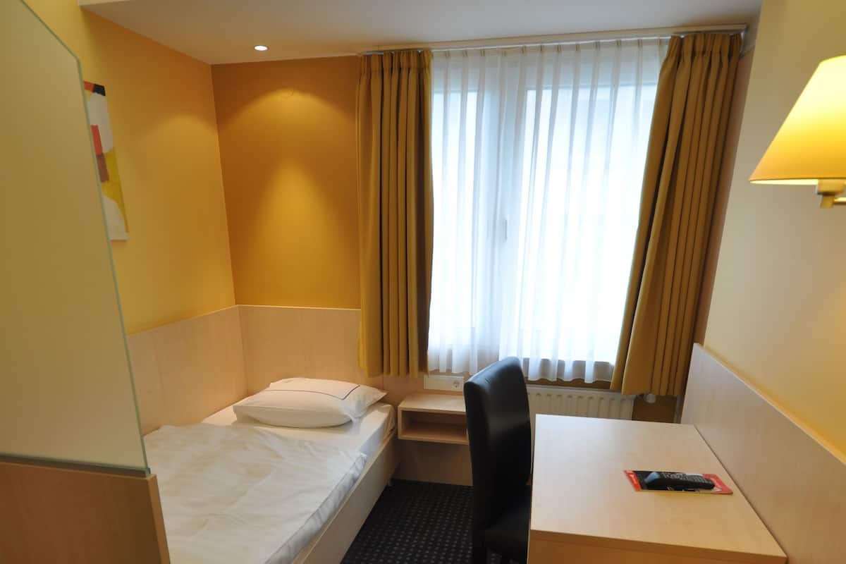 Une chambre d’hôtel avec un lit simple, un bureau, une chaise et une fenêtre avec des rideaux.