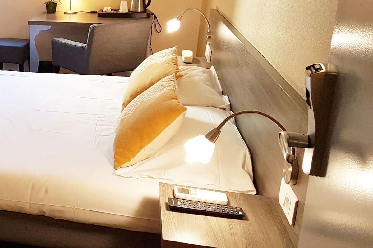 A hotel room with a bed, bedside table, lamp, and a desk with a chair.