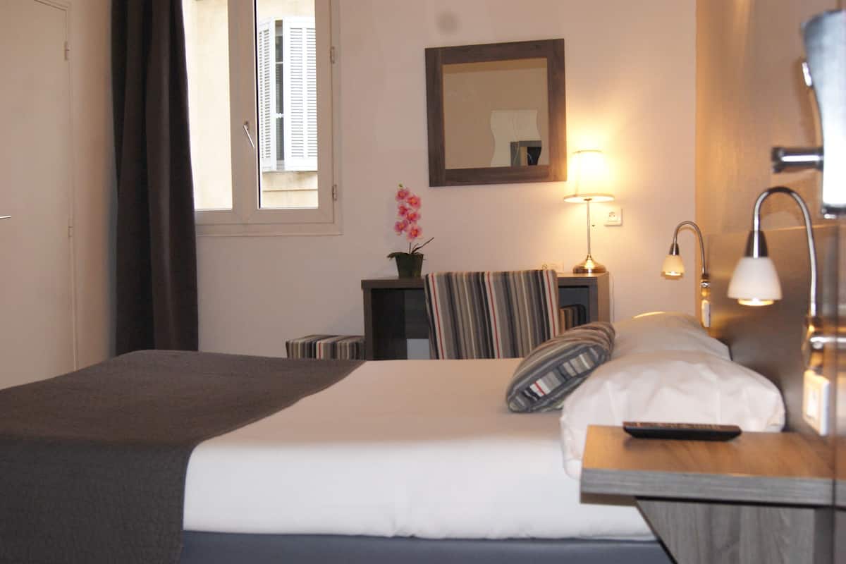 A neatly made bed with a pillow, a bedside table with a lamp, a small table with a plant, and a window with curtains.